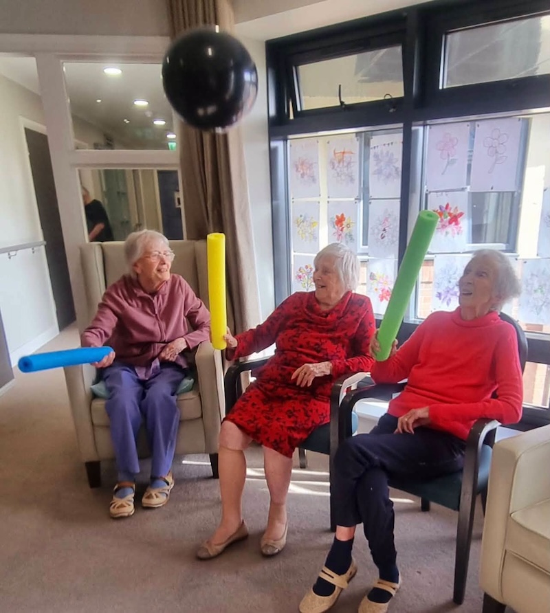3 female residents sat on armchairs holding foam sticks bouncing a balloon