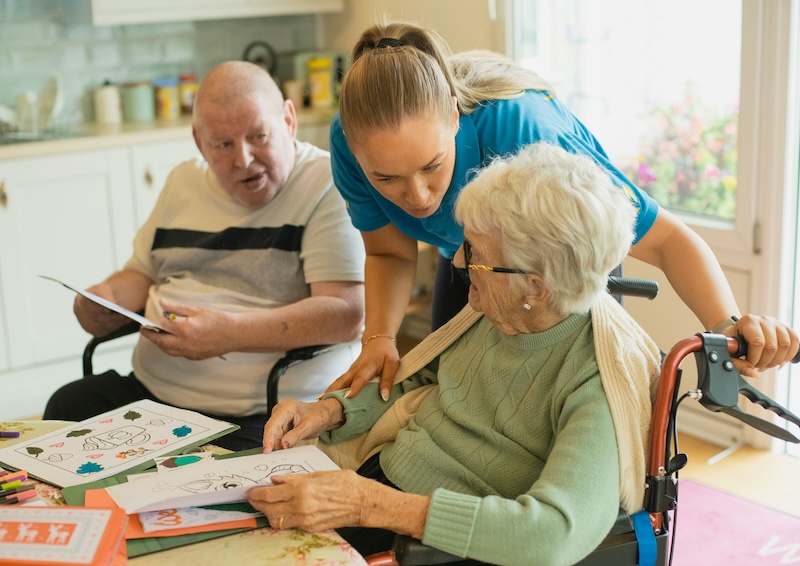 two care home residents sat down doing colouring with a carer supporting them