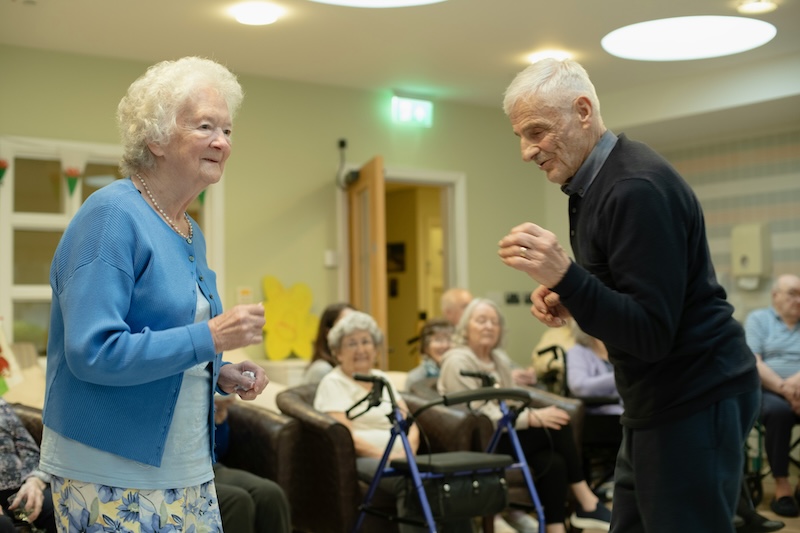 female resident and male resident stood up smiling while dancing in the lounge